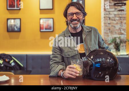 Glücklicher bärtiger reifer Mann mit Helm lächelnd und anblickend Kamera beim Cocktail trinken im Pub Stockfoto