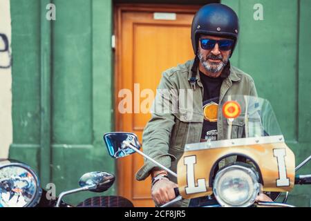 Bärtiger reifer Mann in Helm und Sonnenbrille sitzt auf Custom Motorrad mit vielen Scheinwerfern auf der Stadtstraße Stockfoto
