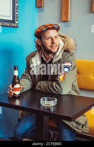 Junger Mann im trendigen Outfit lächelnd und wegblickend Sitzen Sie am Tisch und trinken Sie Bier in der modernen Bar Stockfoto