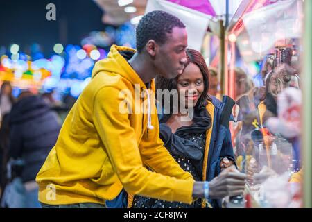 Afroamerikanische Frau lächelt und beobachtet ihren Freund beim Spielen in der Arcade, während sie gemeinsam den Abend auf einem Jahrmarkt verbringt Stockfoto