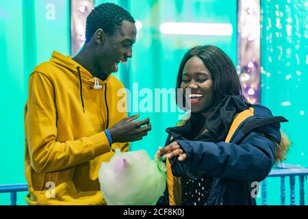 Aufgeregt afroamerikanischen Mann und Frau lachen und essen Zuckerwatte während Datum auf Jahrmarkt Stockfoto