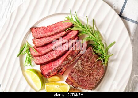 Medium seltenes Steak Rind Striploin in der Pfanne gebraten und in Scheiben geschnitten, serviert auf einem weißen Teller mit Rosmarin und Limetten Keile auf einem weißen Holztisch, , Draufsicht, Clo Stockfoto