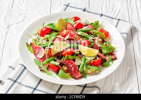 Nahaufnahme auf einem Teller mit Rindersteaksalat, Rucola, Kirschtomaten, grünen Bohnen, zerfetztem Parmesan auf einem weißen Teller mit Limettenkeil Stockfoto