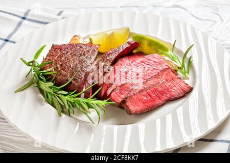 Saftiges, seltenes italienisches Rindfleisch Tagliata Steak in der Pfanne gebraten und in Scheiben geschnitten, serviert auf einem weißen Teller mit Rosmarin und Limettenkeilen auf einem weißen Holztisch, Draufsicht, Stockfoto