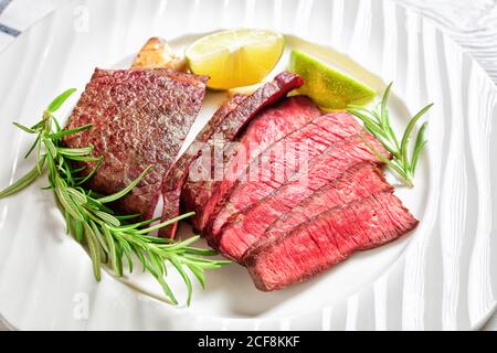 Italienische Tagliata saftiges, seltenes Rindsfilet in der Pfanne gebraten und in Scheiben geschnitten, serviert auf einem weißen Teller mit frischem Rosmarin, geröstetem Knoblauch und Limettenkeilen auf einem Stockfoto