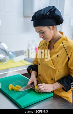 Junge Frau in gelber Uniform und schwarzem Hut schneidet saftige gelbe Früchte auf grünem Schneidebrett, während sie in der Restaurantküche arbeitet Stockfoto