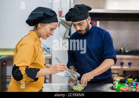 Junge Köchin in gelber Uniform Halteteller und Zange Und männlicher Kollege in dunkelblauer Uniform hält Schüssel mit Essen während der Arbeit zusammen in der Küche und Zubereitung von köstlichen Gericht Stockfoto
