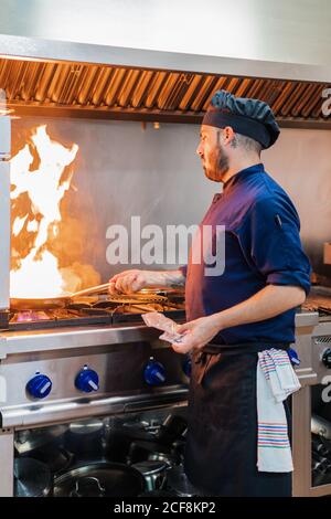 Seitenansicht der männlichen Koch tun Flambe auf dem Essen während Stehen am Gasherd und bereiten Gericht in der professionellen Küche Stockfoto