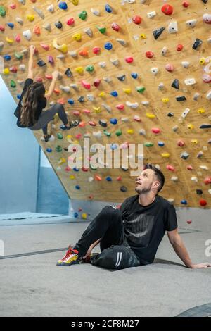 Sportler Mann in Sportkleidung sitzt auf dem Boden und schaut nach oben, während unscharfe anonyme Frau Kletterer Workout an der Wand in der Turnhalle Stockfoto