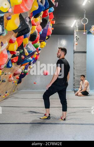 Seitenansicht des nachdenklichen jungen Mannes in Sportswear stehend und Blick auf Kletterwand mit verschwommenem Mann auf der sitzen Hintergrund Stockfoto