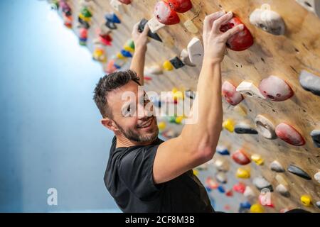 Seitenansicht des starken glücklichen männlichen Athleten, der die Kamera anschaut In Sportbekleidung Klettern an bunten Wand während des Trainings in modern Fitnessraum Stockfoto