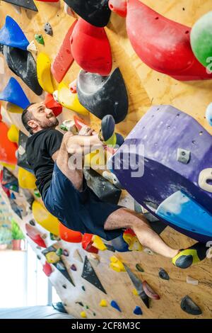 Von bellow starken männlichen Athleten in Sportbekleidung Klettern auf bunt Wand während des Trainings in modernen Kerl Stockfoto