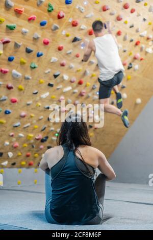 Rückansicht der unkenntlich fokussierten Frau in Sportkleidung, die auf sitzt Boden und beobachten männlich verschwommen anonymen Kletterer an der Wand während Trainieren im Fitnessraum Stockfoto