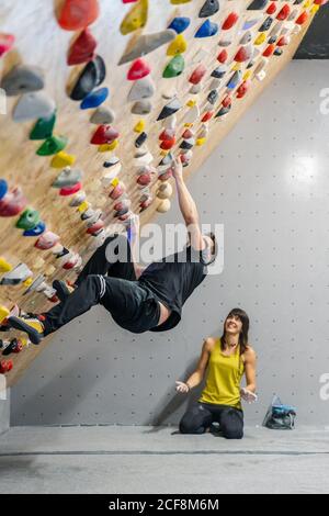 Seitenansicht des männlichen Athleten in aktiver Kleidung Klettern auf Griffe während Bouldertraining mit zufriedenen Weibchen in der Nähe sitzen Fitnessraum Stockfoto