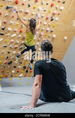 Rückansicht des anonymen männlichen Athleten in Sportkleidung sitzend Der Boden beobachten für verschwommene anonyme Kletterer während des Trainings auf Wand im Fitnessraum Stockfoto