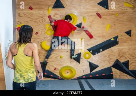 Rückansicht einer Frau in Sportkleidung, die in einem modernen Fitnessstudio steht und den männlichen Teamkletterer an der Wand beobachtet Stockfoto