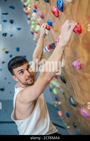 Junger Mann in Sportkleidung, der an der Kletterwand hängt und schaut Während des Bouldertrainings im Fitnessstudio entfernt Stockfoto