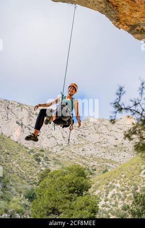 Von unten fröhlich athletische weibliche Alpinistin, die beim Aufhängen wegschaut Auf einer Klippe unter bewölktem Himmel Stockfoto