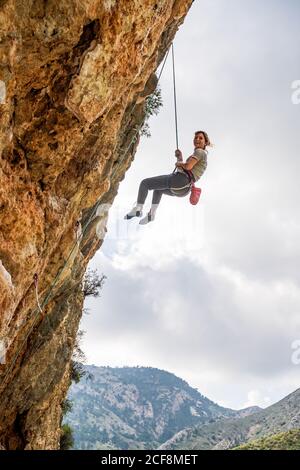 Von unten athletische weibliche Alpinistin hängend auf Klippe unter bewölkt Himmel Stockfoto