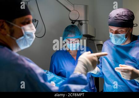 Pensive Chirurg in Uniform Kontrolle Patienten und Entscheidungsfindung während Mitarbeiter mit Kollegen während der Operation im Operationssaal Des modernen Krankenhauses Stockfoto