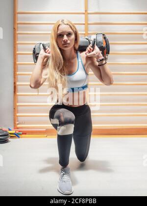 Starke junge Frau in Sportkleidung, die schwere Taschen auf den Schultern hält und die Kamera anschaut, während sie gemeinsam im Fitnessstudio Ausfallschritte macht Stockfoto