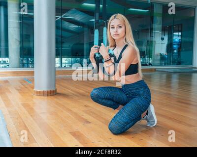 Junge Frau in Sportbekleidung blickt auf die Kamera, während sie trx-Bands beim Training im Fitnessstudio anhält Stockfoto