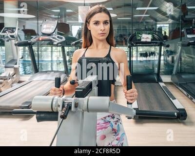 Fokussierte Passform Frau in Sportkleidung, die beim Training auf einem sitzenden Rudergerät im Fitnessstudio auf die Kamera schaut Stockfoto