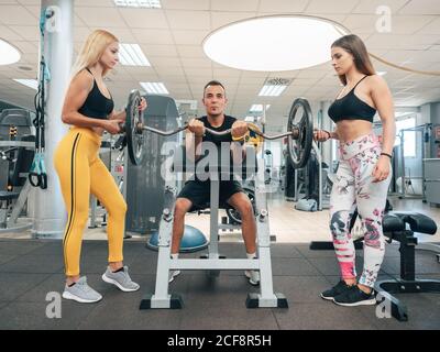 Starke männliche tun Bicep Locken auf der Bank mit jungen schlank Frauen halten Enden der Langhantel in der Turnhalle Stockfoto