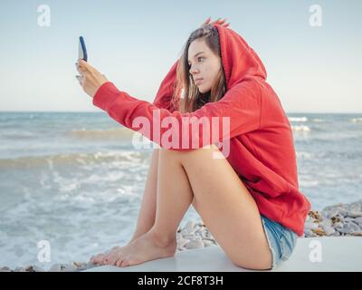 Junge lächelnde Frau mit langen Haaren im roten Hoodie, die Selfie auf dem Smartphone auf dem Surfbrett am steinigen Meer gemacht hat Stockfoto