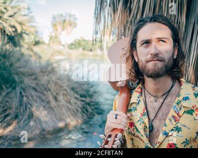 Nachdenklicher bärtiger Hipster, der mit Ukulele im Dschungel unterwegs ist Stockfoto