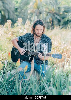 Nachdenklicher bärtiger Hipster, der auf der Wiese sitzt und Gitarre spielt Weg Stockfoto