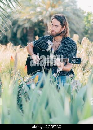 Nachdenklicher bärtiger Hipster, der auf der Wiese sitzt und Gitarre spielt Weg Stockfoto