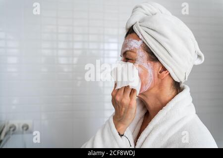 Seitenansicht der Frau in Bademantel und Handtuch Turban stehend Im Badezimmer und Waschen Gesichtsmaske mit nassem Tuch Stockfoto