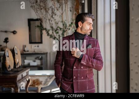 Ruhige pensive Geschäftsmann in karierten Burgunder Jacke mit schwarzem Hemd Und Taschentuch wegschauen und harte Entscheidung treffen, während Stehen gegen verschwommene Vintage-Interieur mit antiken Möbeln und grün Plotted Baum in geräumigen Badezimmer Stockfoto