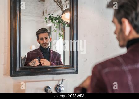 Rückansicht des ruhigen erwachsenen Mannes in eleganter Weinjacke Blick in Spiegel mit Vintage-Holzrahmen und berühren schwarz Taschentuch beim Stehen neben grüner Pflanze und runder Retro Lampe in Licht geräumige Wohnung Stockfoto