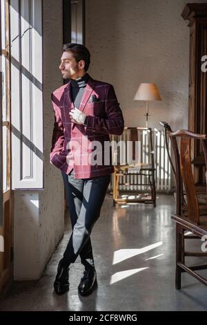 Ruhige pensive Geschäftsmann in karierten Burgunder Jacke mit schwarzem Hemd Und Taschentuch wegschauen und harte Entscheidung treffen, während Stehen gegen verschwommene Vintage-Interieur mit antiken Möbeln und grün Plotted Baum in geräumigen Badezimmer Stockfoto