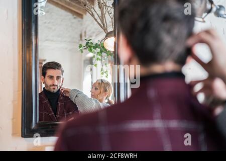 Rückansicht des ruhigen erwachsenen Mannes in eleganter Weinjacke Blick in Spiegel mit Vintage-Holzrahmen und berühren schwarz Taschentuch beim Stehen neben grüner Pflanze und runder Retro Lampe in Licht geräumige Wohnung Stockfoto