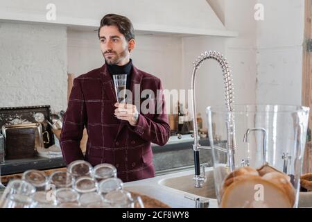 Ruhige nachdenkliche bärtige Unternehmer in eleganten Kleidern mit Glas Champagner schaut weg und betrachtet, während man alleine steht und feiert Leistung gegen vintage Küche Interieur im Landhaus Stockfoto