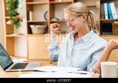 Bild der fröhlichen blonden Frau macht Sieger Geste während der Arbeit Mit Papieren und Laptop am Tisch im Büro Stockfoto