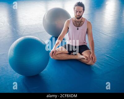 Von oben bärtiger Mann in Sportkleidung mit geschlossenen Augen Und Beine gekreuzt meditieren auf blauem Boden mit Gymnastikbällen Im Studio Stockfoto