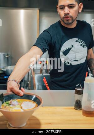 Junger Mann in schwarzem T-Shirt setzen auf Tisch Schüsseln von Frisch gekochtes japanisches Gericht namens Ramen Stockfoto