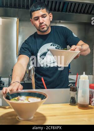 Junger Mann in schwarzem T-Shirt setzen auf Tisch Schüsseln von Frisch gekochtes japanisches Gericht namens Ramen Stockfoto