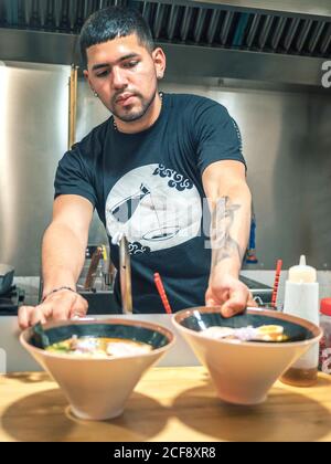 Junger Mann in schwarzem T-Shirt setzen auf Tisch Schüsseln von Frisch gekochtes japanisches Gericht namens Ramen Stockfoto