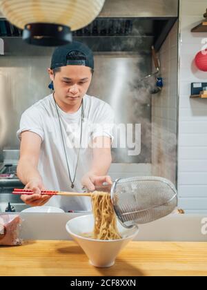 Junger Mann, der heiße Nudeln mit Essstäbchen in die Schüssel gibt Kochen japanische Gerichte in der Küche Stockfoto