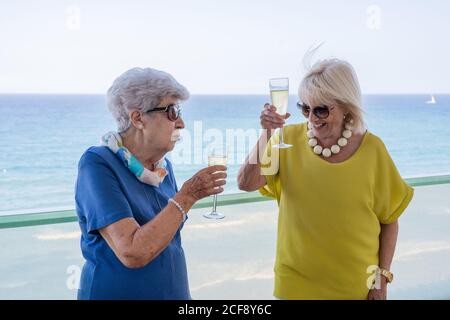 Glückliche reife Frauen in eleganten Outfits sagen Toast und genießen Wein auf dem Hotelbalkon in der Nähe des Meeres im Resort Stockfoto