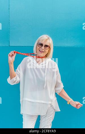Porträt einer schönen, stilvollen weißhaarigen älteren Frau mit Sonnenbrille in weißem Hemd mit Kapuze auf dem Kopf und Korallenkugeln auf blauem Wandhintergrund Stockfoto