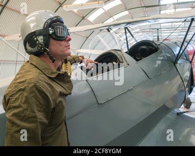 Seitenansicht des männlichen Piloten in der Ausrüstung, die neben dem Vintage steht Doppeldecker im hellen Hangar Stockfoto