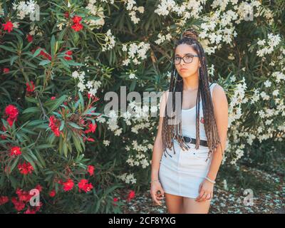 Schöne junge Frau in Brille Sommerkleidung und mit Dreadlocks stehen in der Nähe blühender Bäume und Blick auf die Kamera Stockfoto