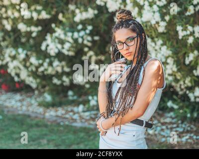 Stilvolle junge Frau trägt Brille Dreadlocks und Sommerkleidung mit Smartphone und Blick auf die Kamera Stockfoto