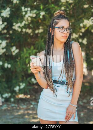 Stilvolle junge Frau trägt Brillen Dreadlocks und Sommerkleidung mit Smartphone und schaut weg Stockfoto